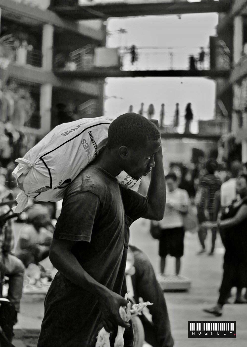 Black and white documentary photograph of a man carrying a sack through a market